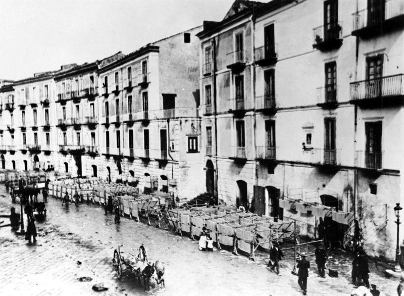 A image taken from the second floor of a building shows the above view of an Italian street where rows of drying racks line buildings with noodles hanging from them drying in the sea breeze.