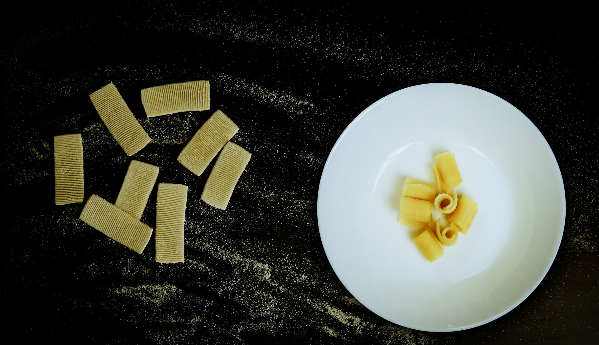 An photograph shows a white plate with cooked rigatoni, on the side in contrast are dried, flat pastas similar in shape
