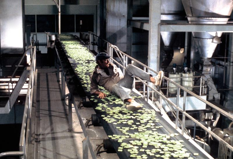 A man slides down a conveyor belt in a factory surrounded by small green tags. 