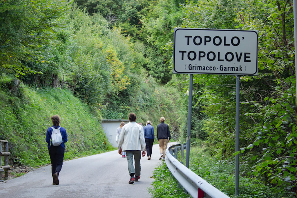 A group of people walk in the road into the village of Topolov, in a densely forested area.