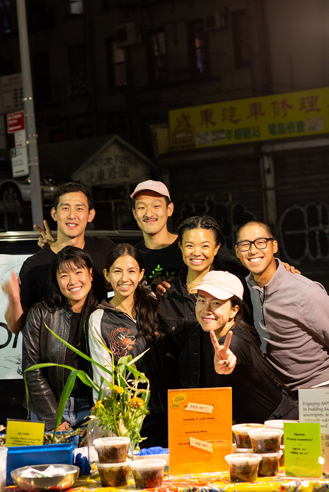 A group of seven individuals of Asian descent stand around a table vending food with their arms around one another.