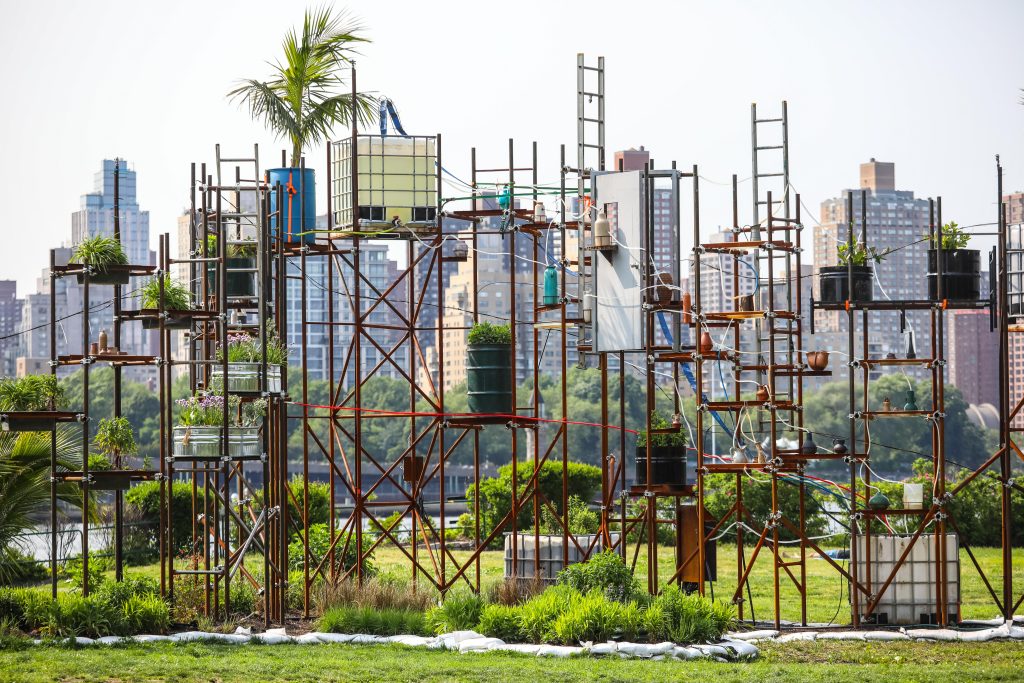 A structure constructed with scaffolding, ladders, and tubs of water and planters sits outside on a grass field. The skyline of New York City is in the backdrop.