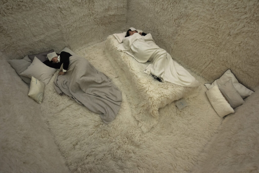 Two individuals lie coddled in blankets with their eyes covered in a room completely covered on all surfaces with a shaggy white fur carpeting.