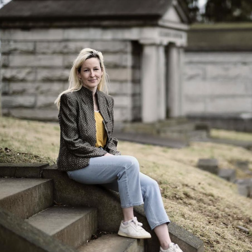A white woman sits on concrete steps in a khaki jacket, blue jeans and white sneakers.