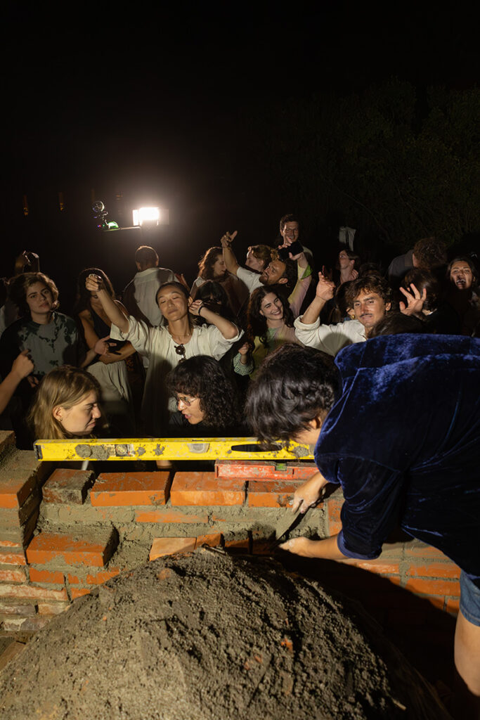A group of people gather around a brick-laying project at night. 