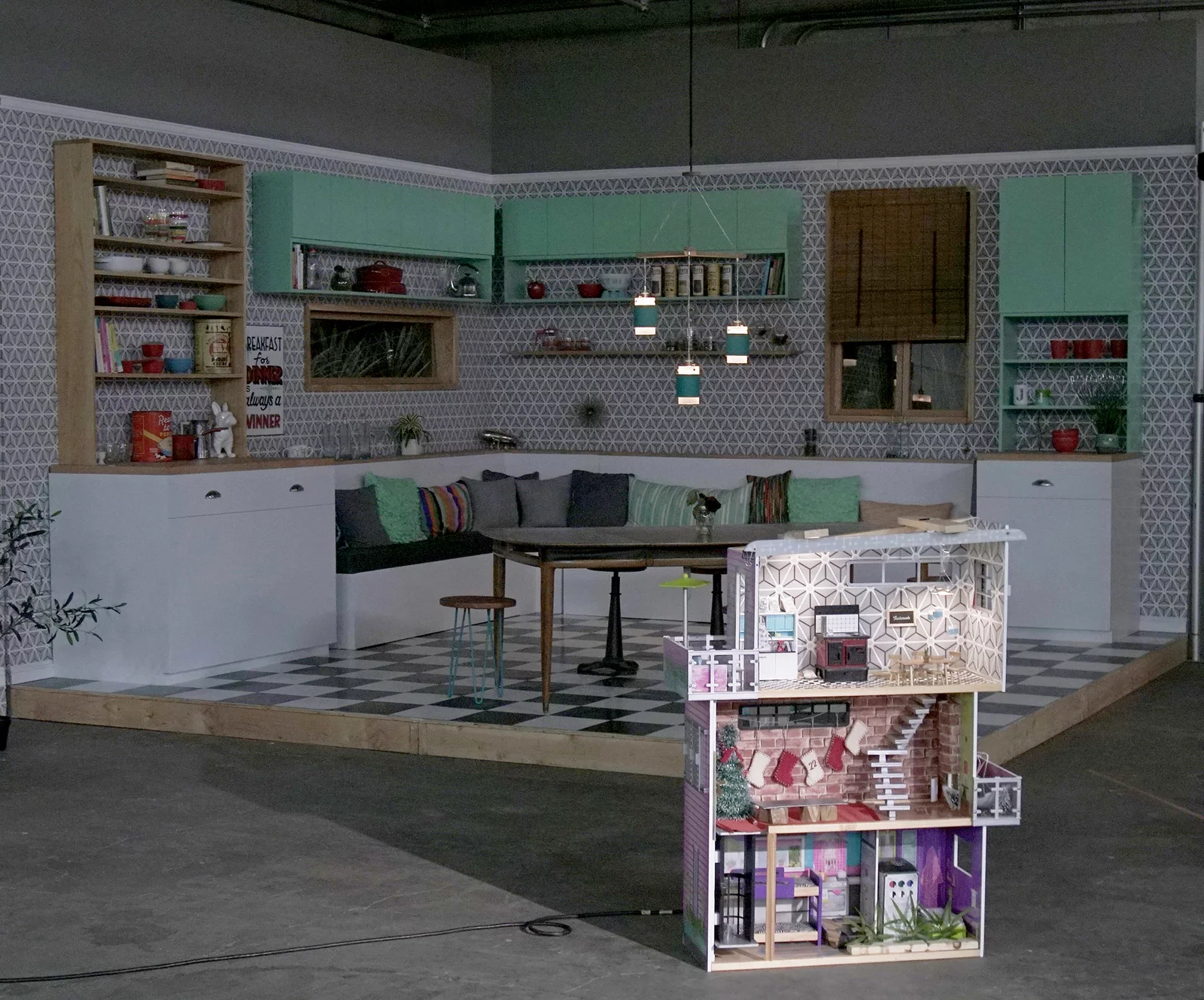 On a soundstage, a kitchen set is in the back whereas in the foreground a dollhouse sits off the soundstage.