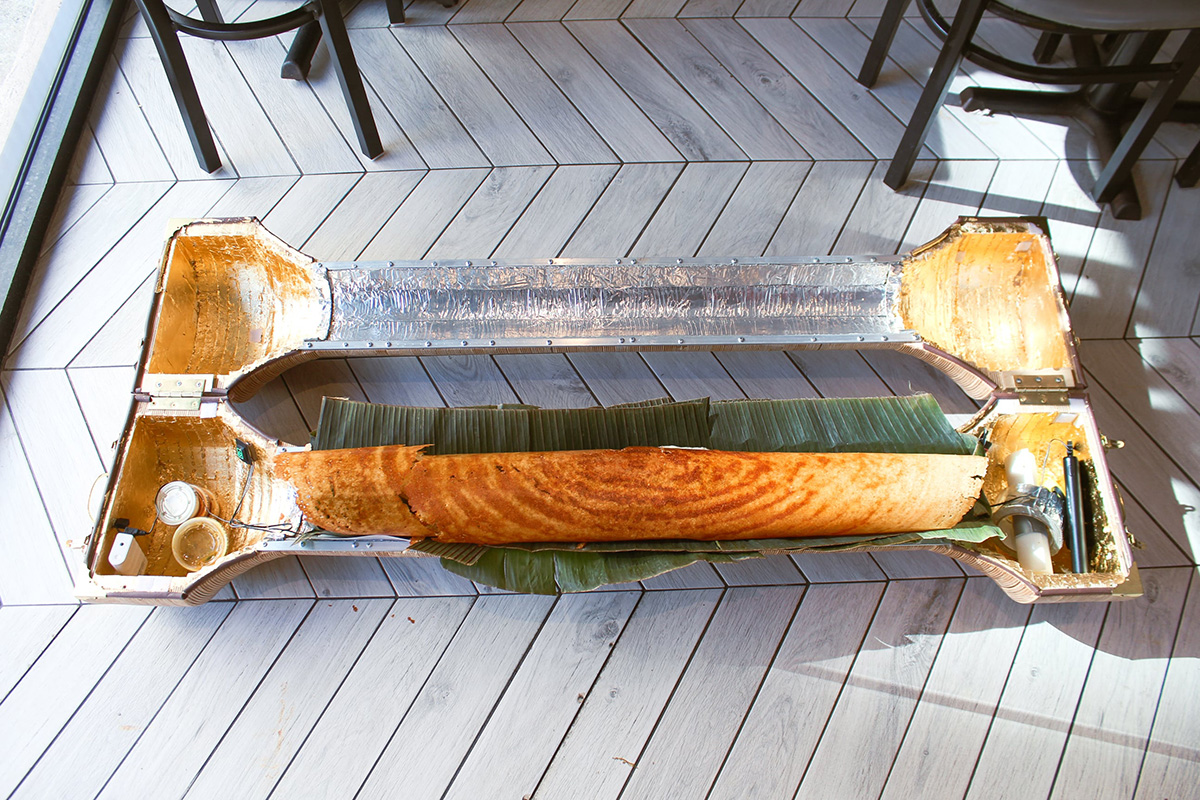 A wooden cylindrical Dosa picnic basket sits open on the floor. Inside it hold a Dosa wrapped in banana leaves.