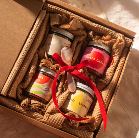 A cardboard box opened to show four jars of condiment nestled in burlap with a red bow on top. 