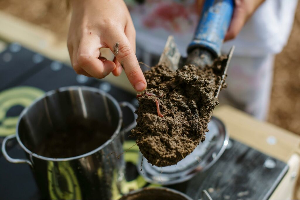 A child's hand points at a shovel holding a load of compost where small worms wriggle about.