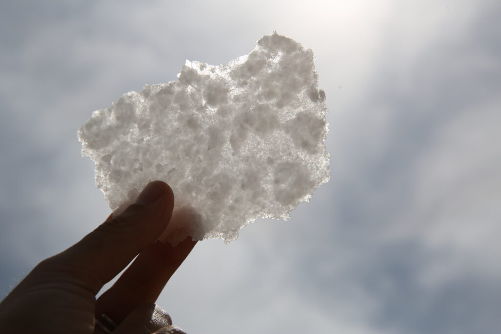 A large flake of salt is held up to the sunlight against the sky. 