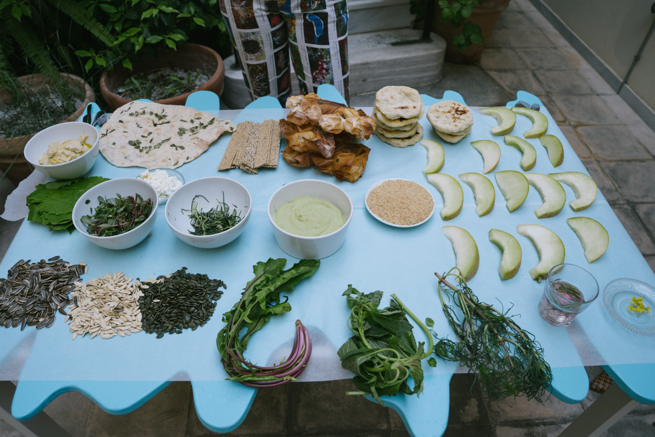 A blue wavy table features a crudite of halophytic plants and vegetables, dips, and round discs of bread.