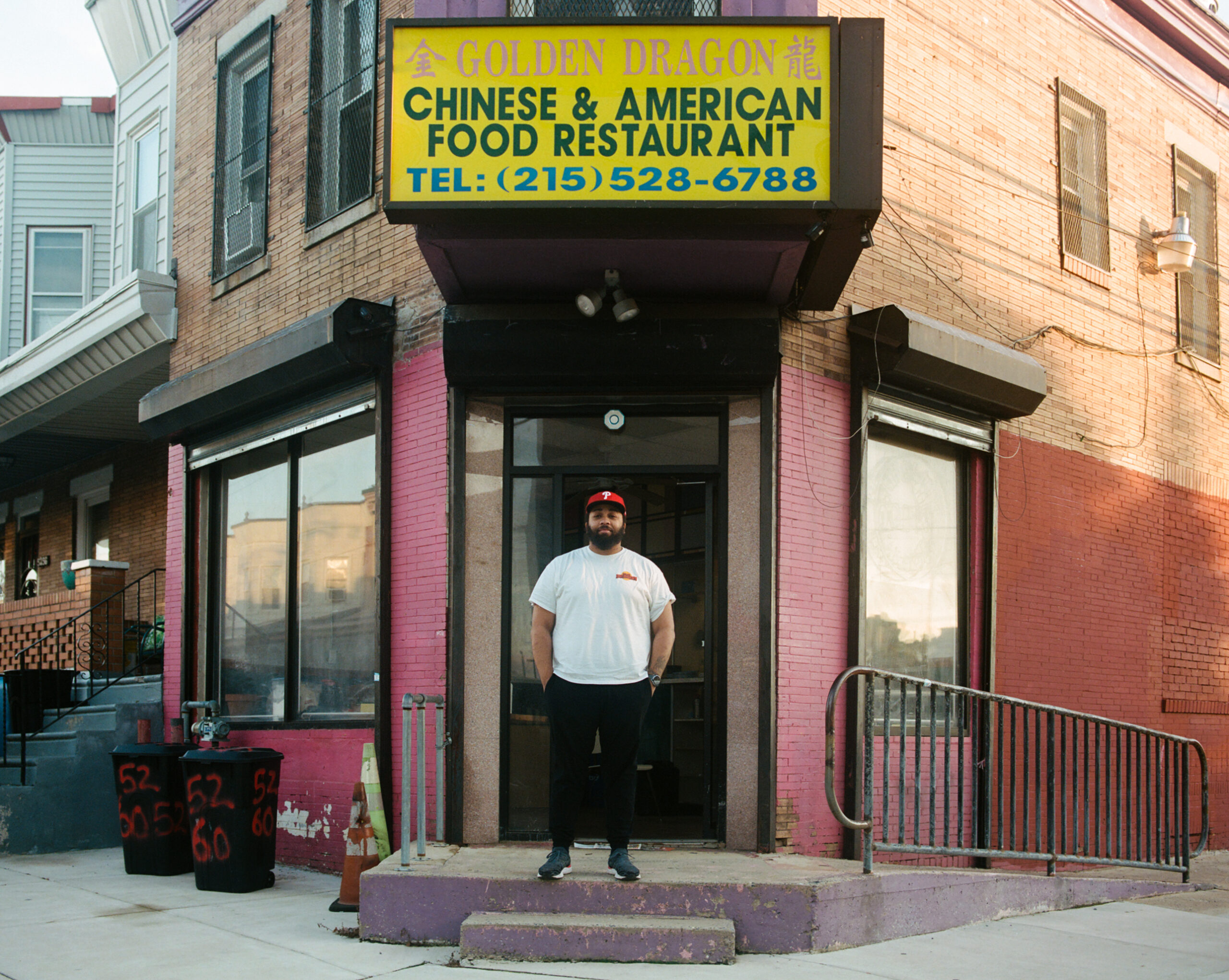 A black man in a white shirt and red cap stands in front of a corner storefront with his hands in his pocket. A yellow sign above him says Golden Dragon Chinese & American Food Restaurant.