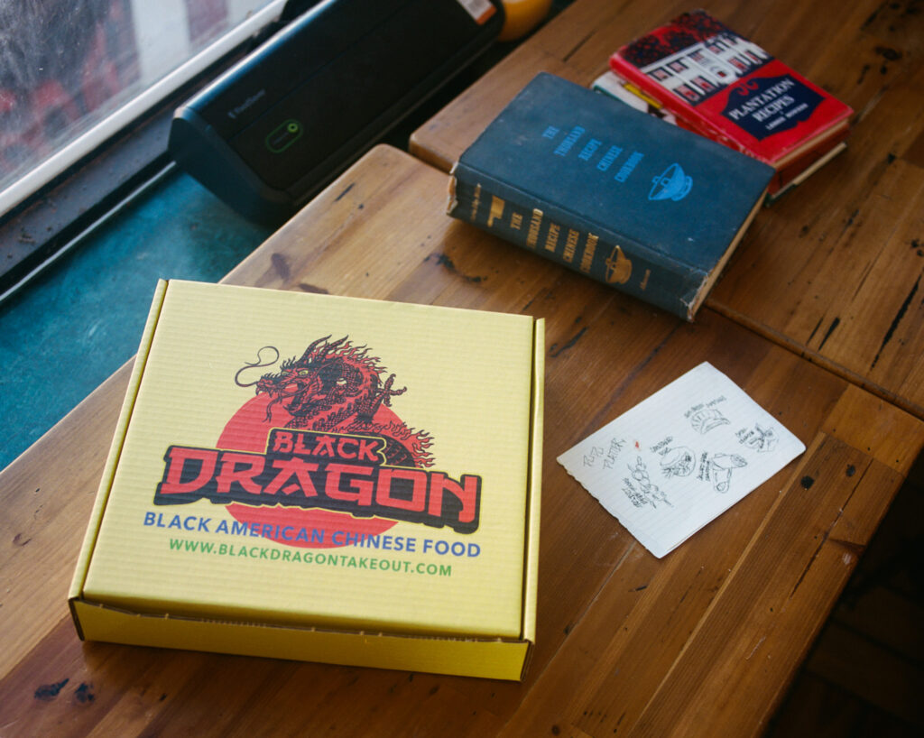 A yellow cardboard takeout box with a dragon on it sits on a wooden table next to two books and a sketch of dishes.