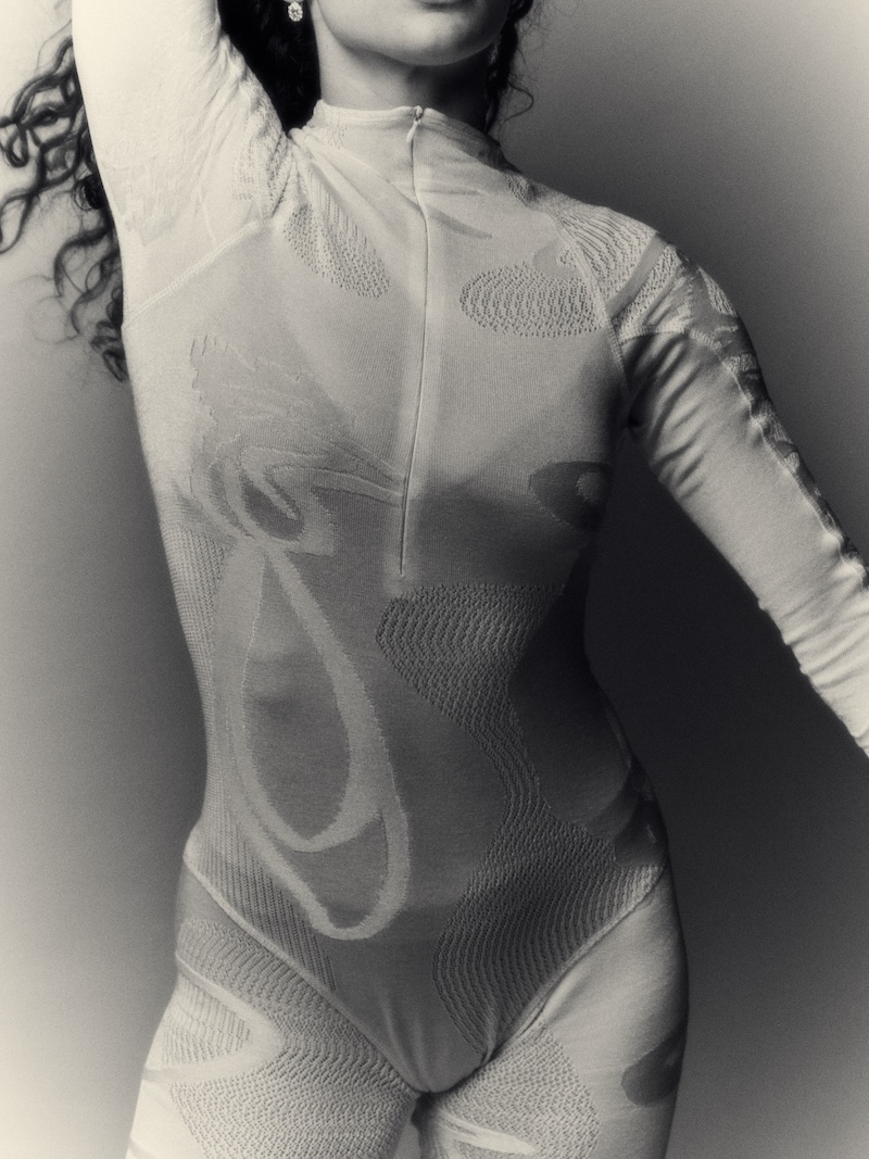A black and white photo of a woman's torso is shown wearing a white knitted body suit and tights. Her arms are caught in motion.