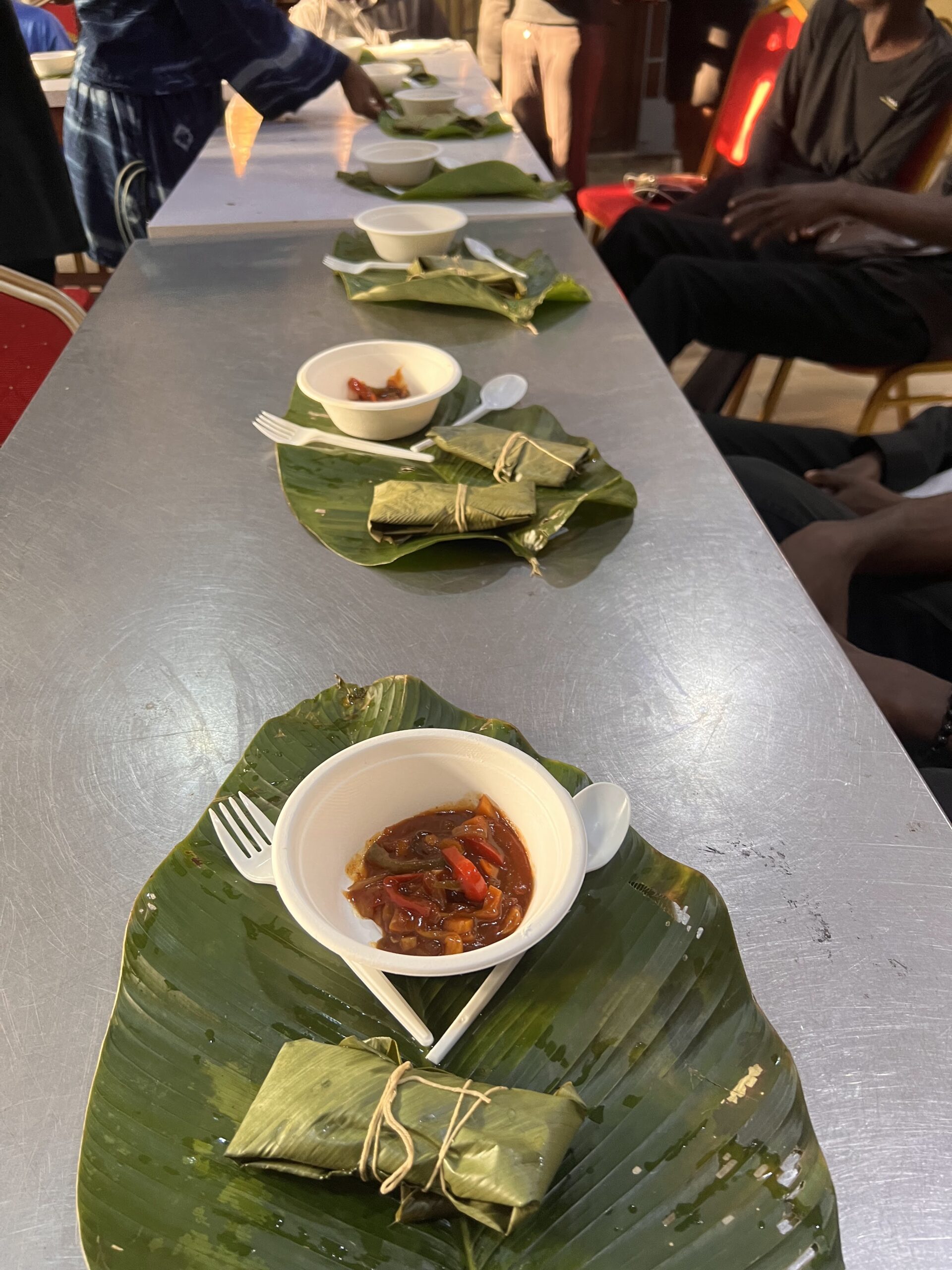 A long table with set with banana leaves, with a wrapped cinavu on top as well as sauce and a fork and spoon.