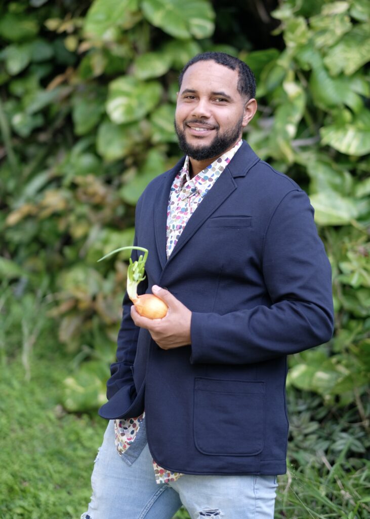 A man in a navy suit holds a sprouting onion in his hand while looking at the camera.