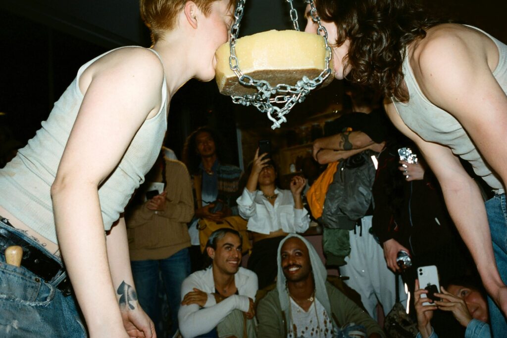 Two individuals wearing white tank tops take bites out of a cheese wheel hung from the ceiling by a chain. An audience looks on in the background.