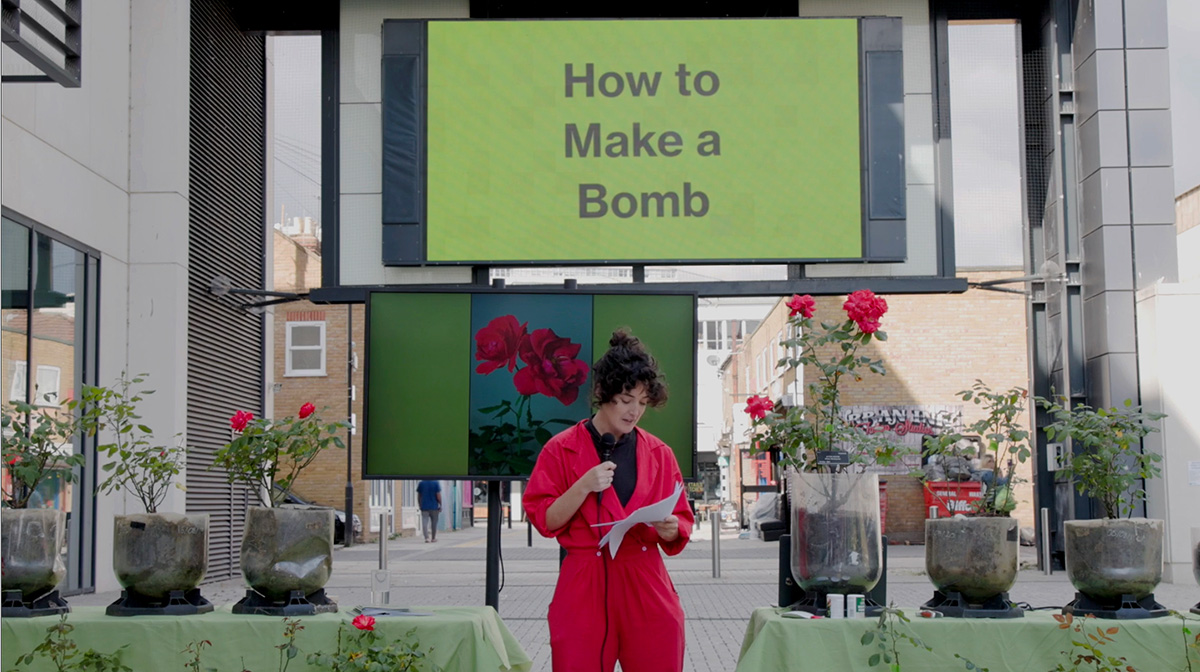 A woman wearing a red jumpsuit stands in front of various rose bushes speaking into a microphone. Behind her is a screen with a green background that says How to Make a Bomb