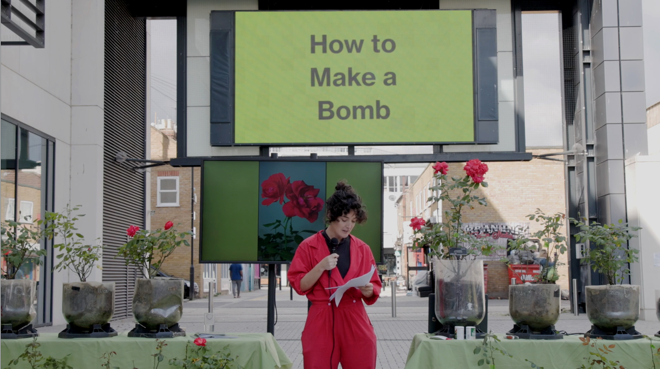 A woman wearing a red jumpsuit stands in front of various rose bushes speaking into a microphone. Behind her is a screen with a green background that says How to Make a Bomb
