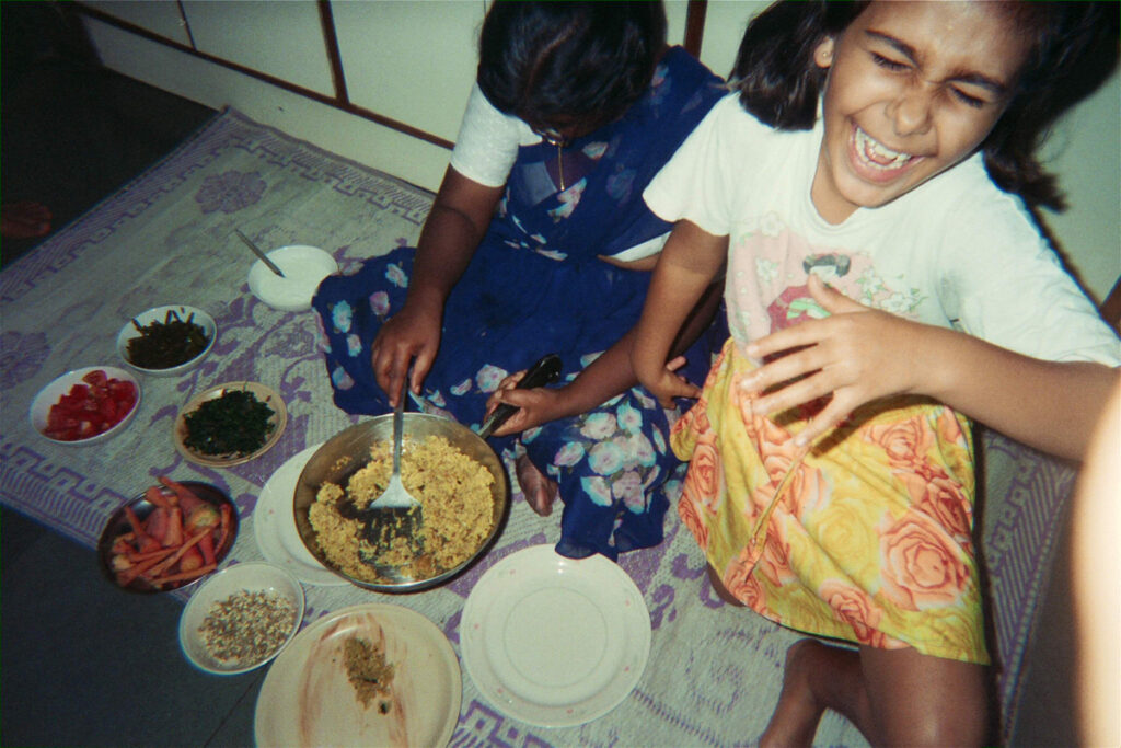 One boy and one girl sit on the floor where they have plates with different foods laid out before them. The boy digs in with a large spoon. The girl laughs facing the camera.