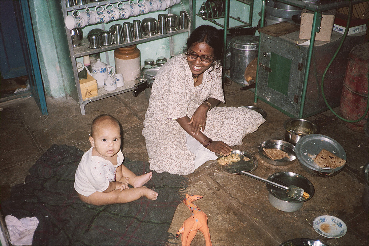 A baby and a woman sit on the floor surrounded by silver plates and vessels filled with food. They are in a kitchen. The woman smiles down at the baby.