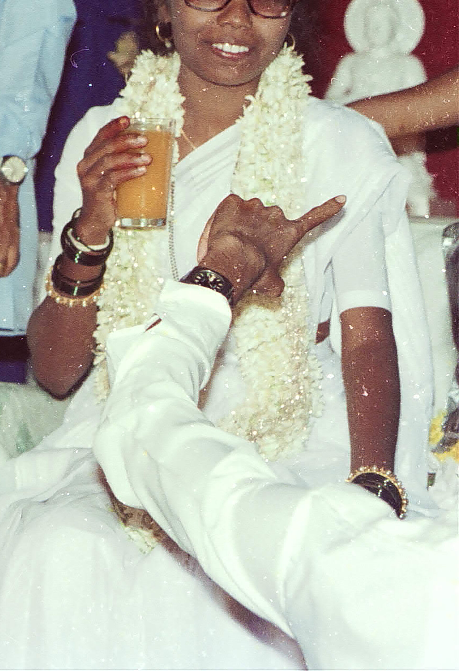 A woman wearing a white sari and a garland of white flowers holds a glass with an orange beverage smiling, a man whose body is off screen's arm points toward her.