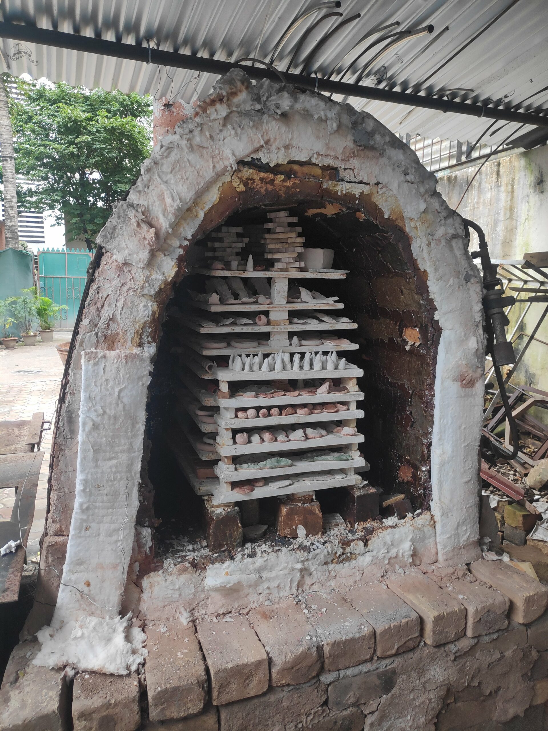 A brick kiln sits outdoors under a corrugated tin roof. It is filled with small ceramic creations.