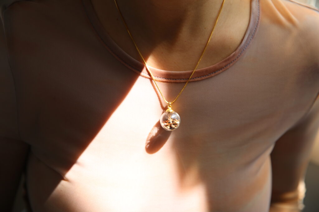 A woman wearing a tan sheer mesh top and a gold necklace with a glass sphere attached as a pendant, seeds are inside the sphere. 