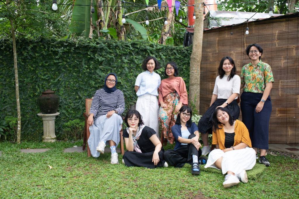 Eight women stand in a yard, some squatting some standing. All wearing coloroful clothing with a tree int he background.