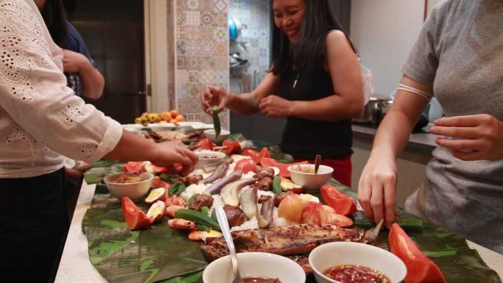 A table with women smiling around it is set with banana leaves and various dishes.