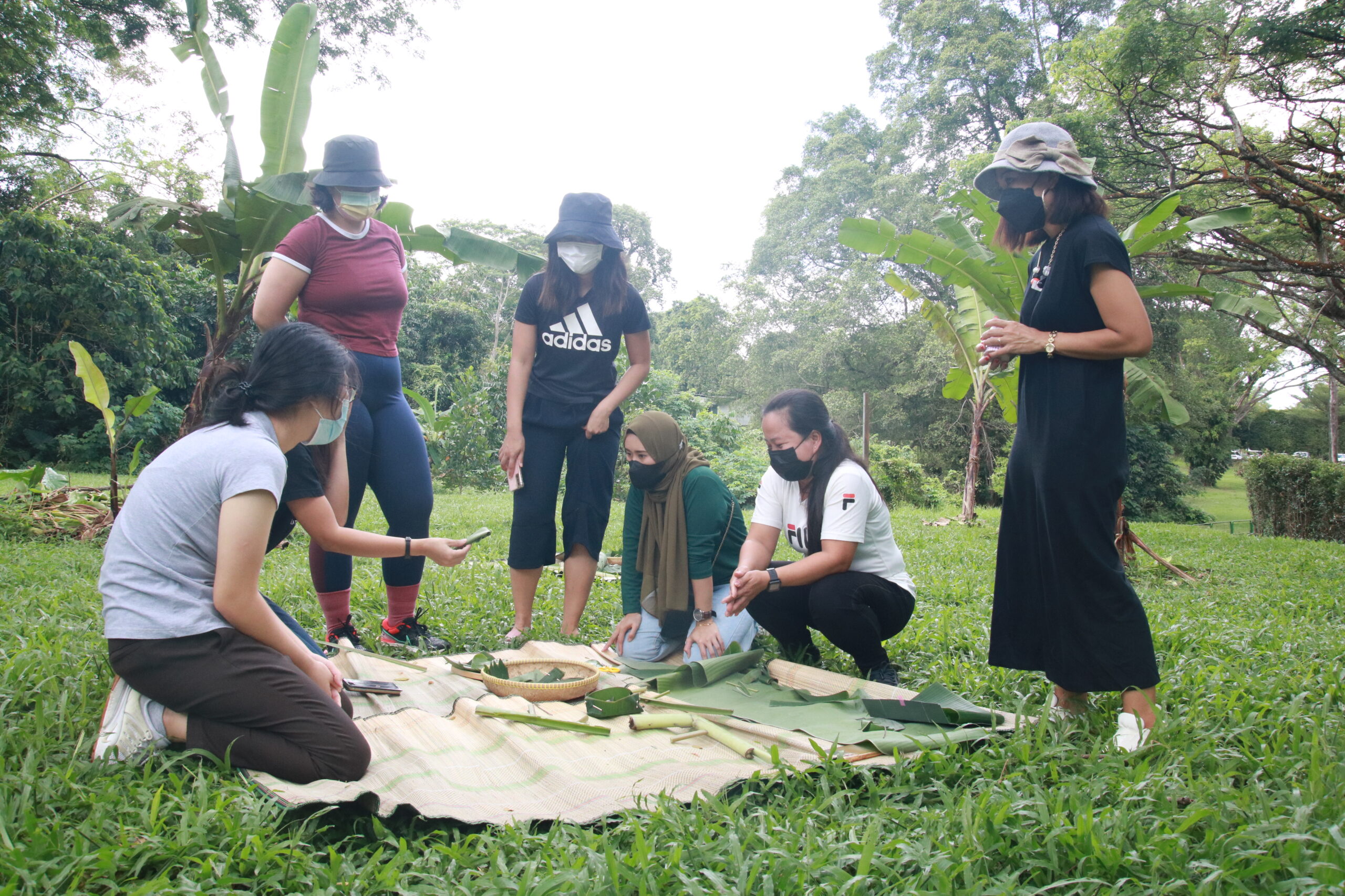 Six women crouch around a blanket in a verdant park placed on grace. On this blanket are banana leaves and baskets.