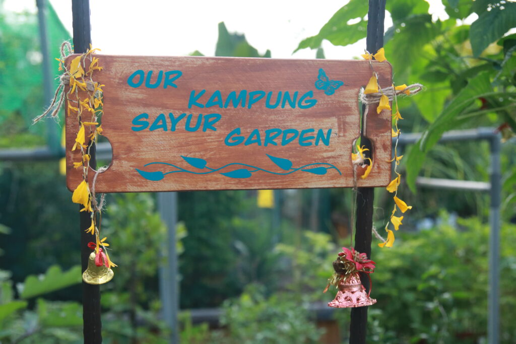 A wooden sign that says Our Kampung Group Garden. Bells and flowers are tied to the wooden sign.