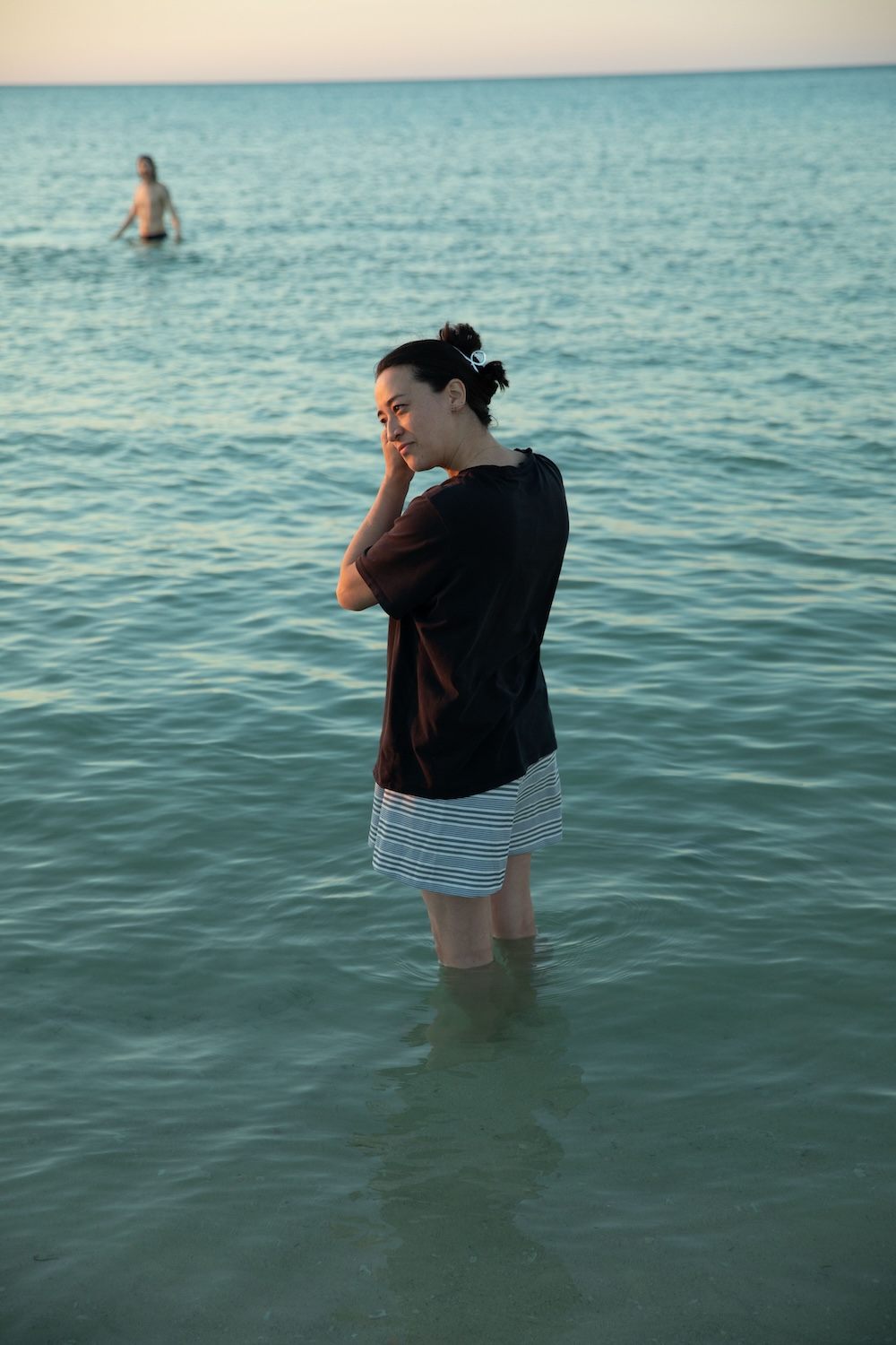 An image of a woman wearing shorts and a tshirt who has waded knee deep into the ocean. She fiddles with an earring.